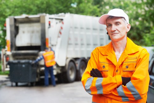 A male waste removal worker wearing an orange high-visibility jacket with reflective stripes and a white cap stands outdoors on a driveway or street in front of a large industrial waste collection vehicle, which is in the process of emptying a black rubbish bin into its open rear hopper. The vehicle is predominantly white with red and yellow safety markings along the edges. The background features green trees and foliage, indicating a residential or urban environment in the vicinity of Pimlico, Westminster. The worker has a neutral expression, arms crossed, and the scene is well-lit, suggesting daytime. The setting, attire, and equipment reflect typical rubbish clearance activities undertaken by Pimlico House Clearance services, with a focus on domestic waste collection in central London areas, including postcode regions associated with the vicinity. The image captures the precise moment of waste transfer, highlighting the professional process of rubbish removal within a local urban context.