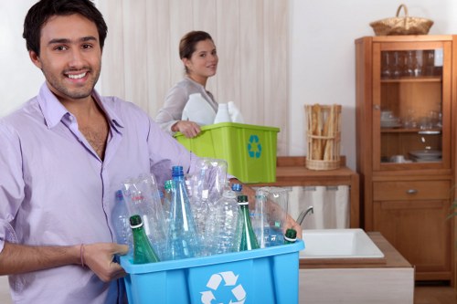 In a domestic kitchen setting, a man with a cheerful expression holds a blue recycling bin filled with clear plastic bottles, some with green tint, and metal caps, positioned in the foreground on the left side of the image. Behind him, a woman with a neutral expression faces to the right, standing near a green recycling container with a recycling symbol on it, placed on a light wooden surface. The kitchen features a white sink and a wooden hutch with glass doors, displaying various dishes and glassware, on the right side of the background. Light-colored curtains hang in the background, and the overall environment suggests a clean, organized space conducive for waste separation. The scene emphasizes household waste management and recycling activities, aligning with rubbish removal services offered by Pimlico House Clearance in London, capturing a typical home recycling process context within a residential setting, possibly in central London or nearby suburban areas.