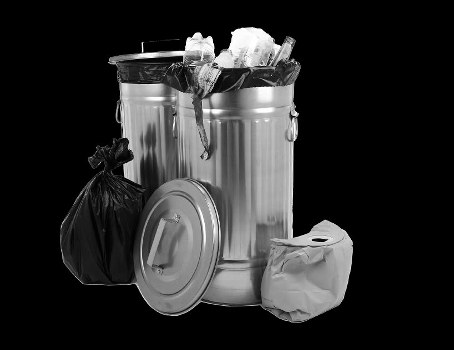 A cylindrical metal rubbish bin with a partially open lid atop it, containing visible crumpled paper, plastic bottles, and other miscellaneous waste items. Leaning against the bin is a tied black plastic trash bag, with a round metal lid resting on the ground beside it. To the right of the bin, there is a crumpled paper waste bag. The scene is set against a plain black background, emphasizing the metallic surface of the bin and the various discarded items, which represent typical household or commercial rubbish. The overall imagery conveys waste collection and disposal, relevant to rubbish removal services such as those offered by Pimlico House Clearance in London, with a focus on general waste segregation and clearance within a local context.
