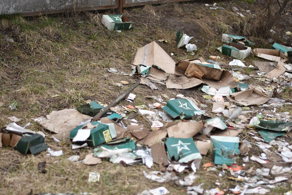 An overflowing outdoor rubbish collection point situated on a paved area, featuring multiple black, green, and red waste bins filled with mixed household waste, including paper, cardboard boxes, plast