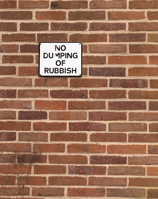 A close-up image of a black and white metal sign mounted on a red brick wall. The sign reads 'NO DUMPING OF RUBBISH' in bold uppercase letters, with the word 'DUMPING' partially obscured or faded. The